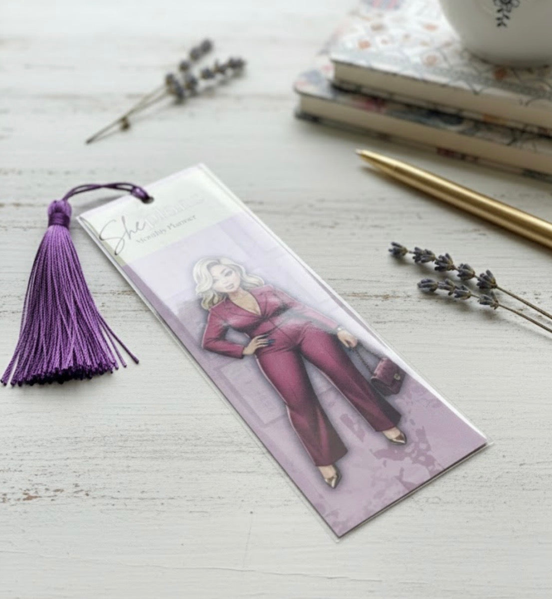 Bookmark with illustration of a person in a burgundy outfit and purple tassel on a light wooden surface.