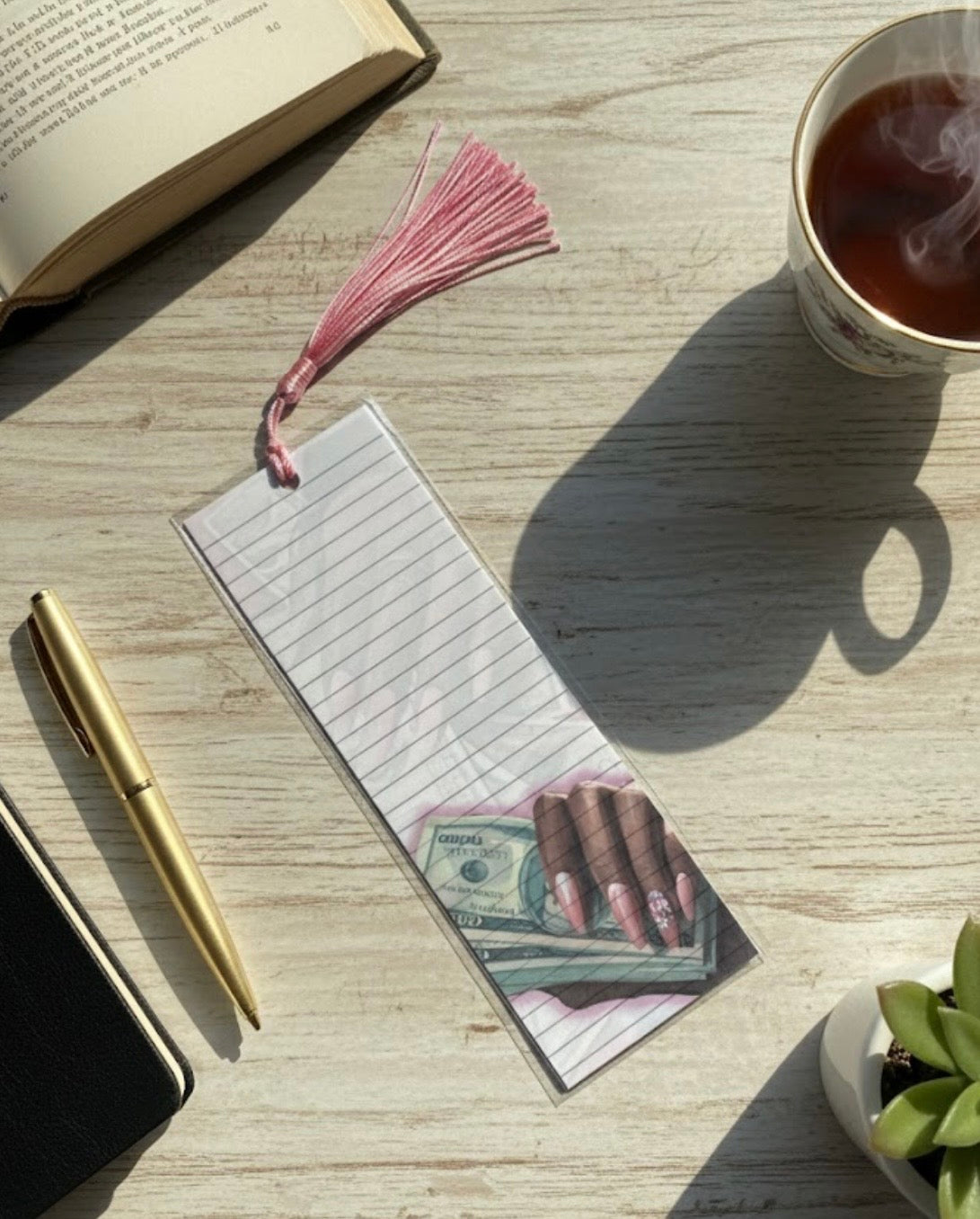 Bookmark with money design on a wooden surface with a book, pen, and cup of tea.