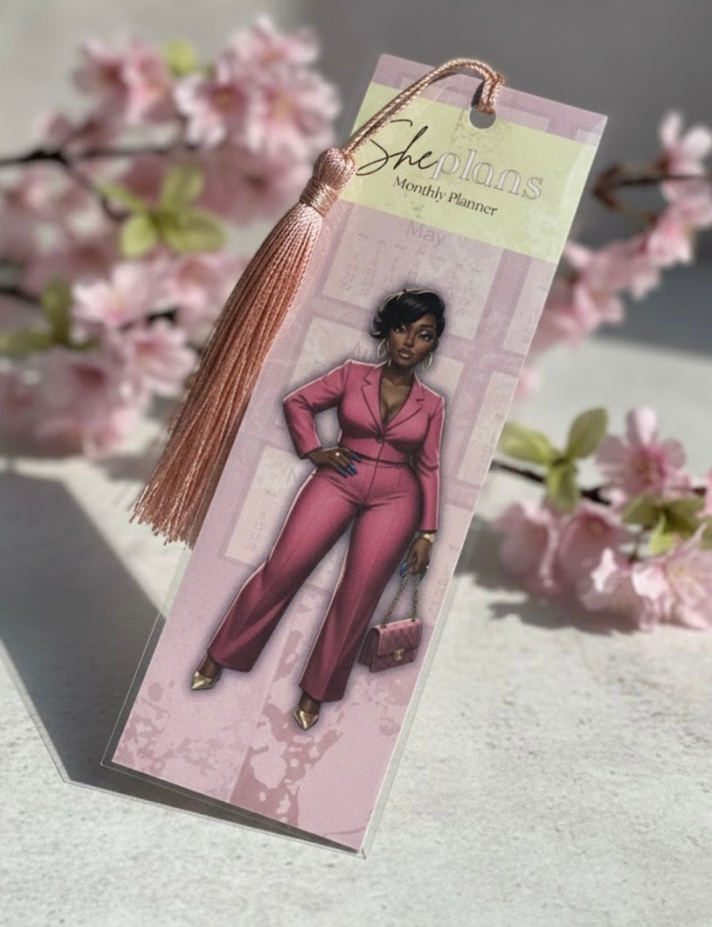 Bookmark with a person in a pink outfit and tassel, placed on a surface with cherry blossoms in the background.