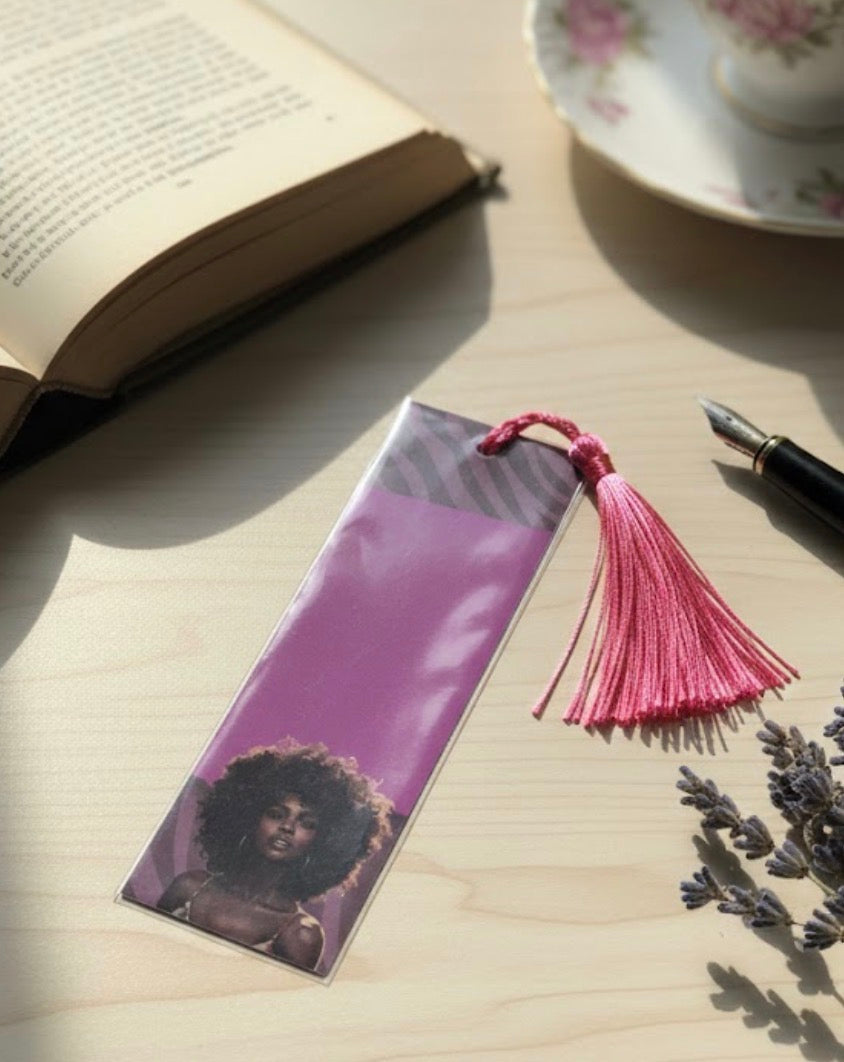 Bookmark with a pink tassel on a wooden surface with a book and tea cup in the background