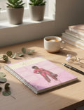 Planner with a pink cover featuring an illustration of a woman, placed on a wooden desk with a cup of coffee and books in the background.