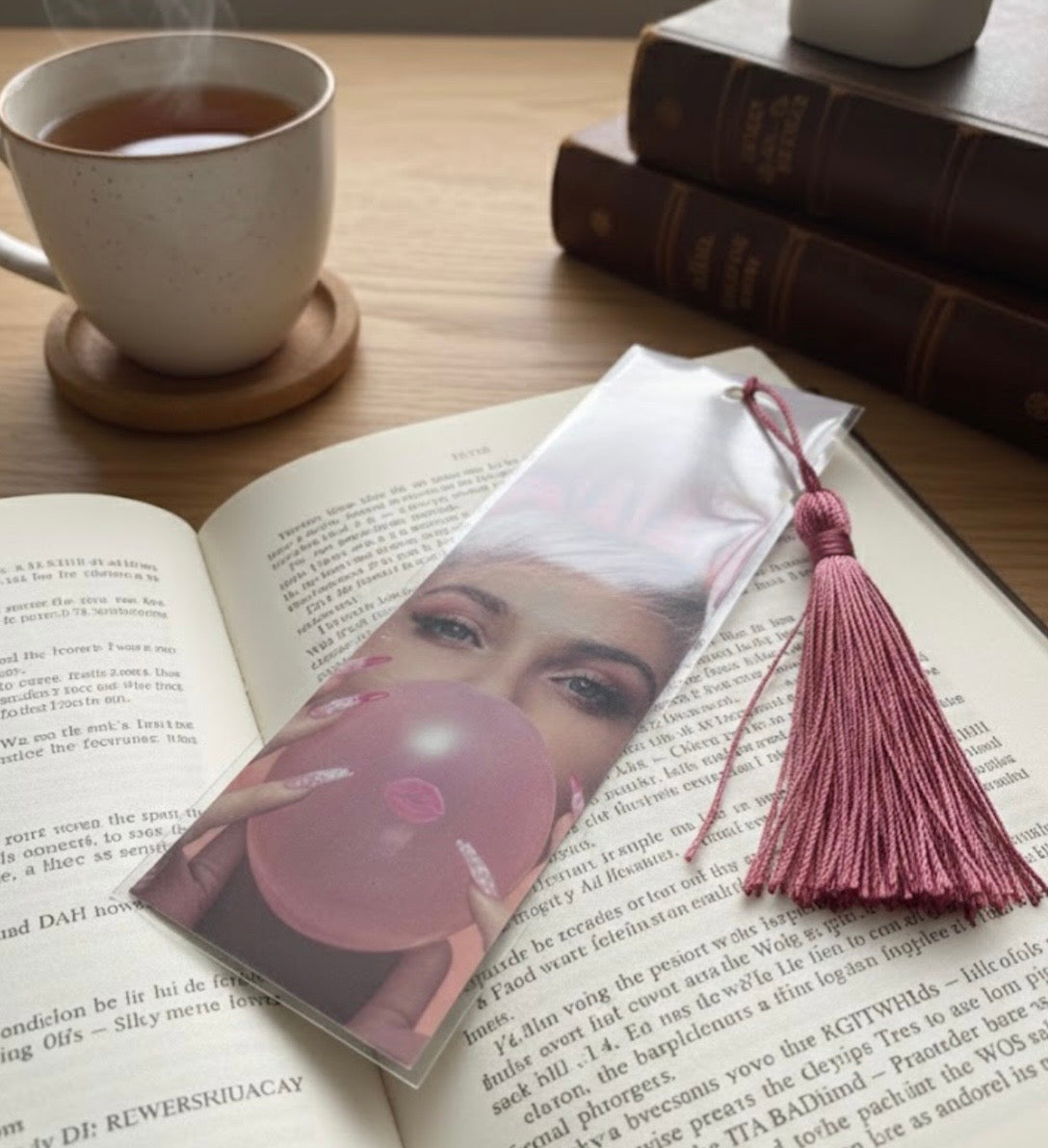 Open book with a bookmark featuring a woman blowing a bubble, next to a steaming cup of tea on a wooden table.
